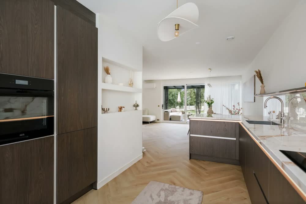 Hoge kastenwand in donker houtfineer met een inbouwoven, open nissen in de wand en een doorkijk naar de woonkamer en de tuin
