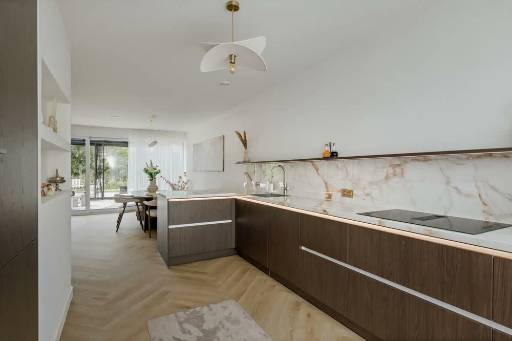 L-vormige keuken in donker houtfineer met een schiereiland, een lichtlijn onder het werkblad en een achterwand in marmerlook tot aan de wandplank