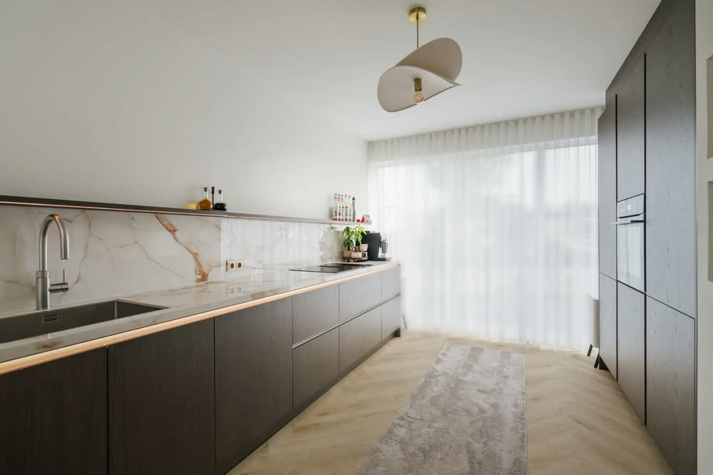 Lange keukenwand in donker fineer met spoelbak en kookplaat, rechts de hoge kastenwand en vitrage voor de pui