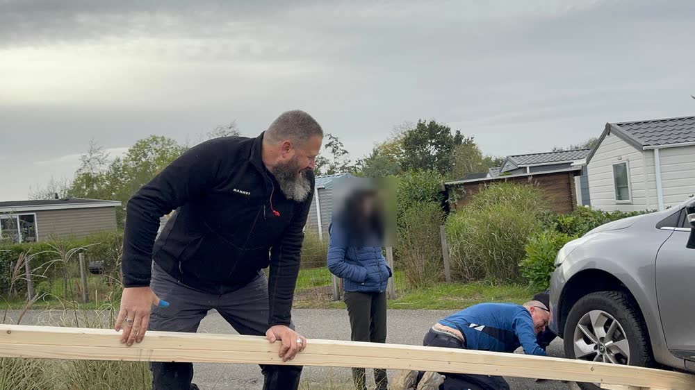 Een van onze vakmensen legt balken klaar terwijl een collega onder de auto kijkt; de bestuurster staat erbij