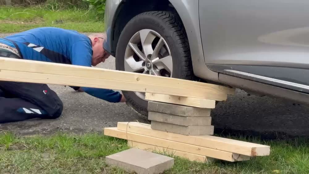 De auto is met stoeptegels en balken onderstopt; een balk dient als hefboom terwijl een collega onder de auto werkt
