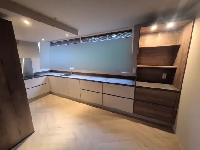 U-keuken van zo’n tien meter onder een breed raam, met zandkleurige fronten en een open nis in donker eiken